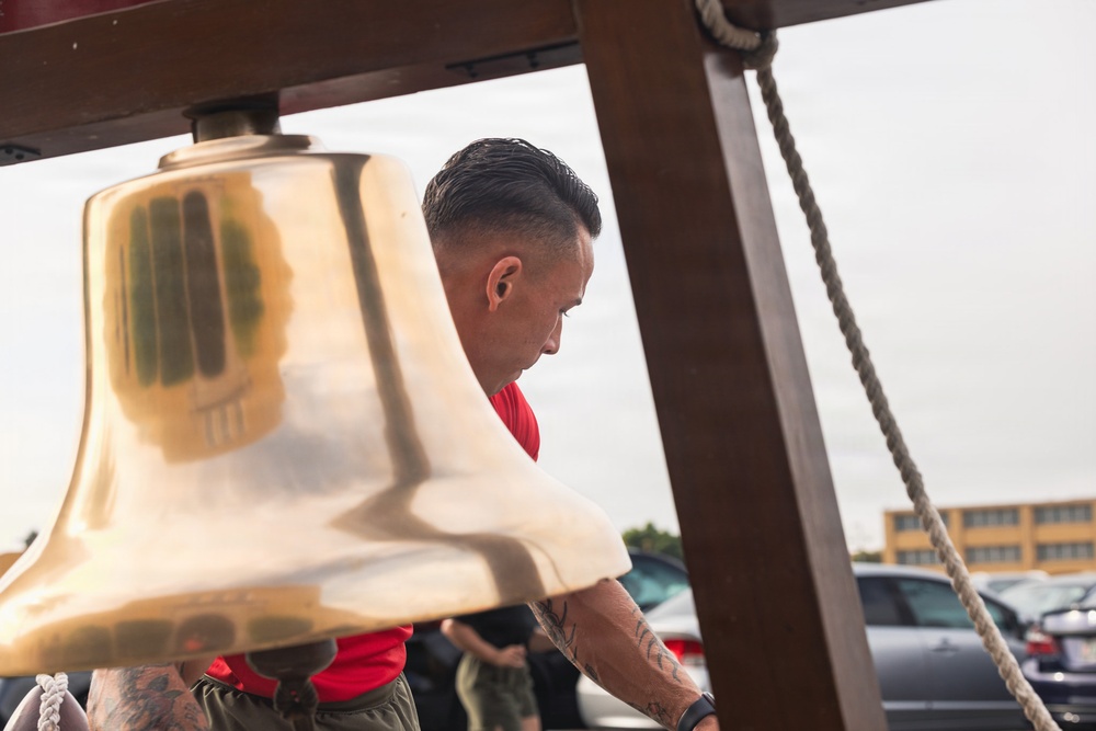 A man with a military-style haircut and visible tattoos on his forearm leans toward a large brass bell mounted on a wooden frame. The setting appears to be an outdoor area with a parking lot and building visible in the background. The scene suggests a ceremonial or commemorative event, possibly military or first responder related.