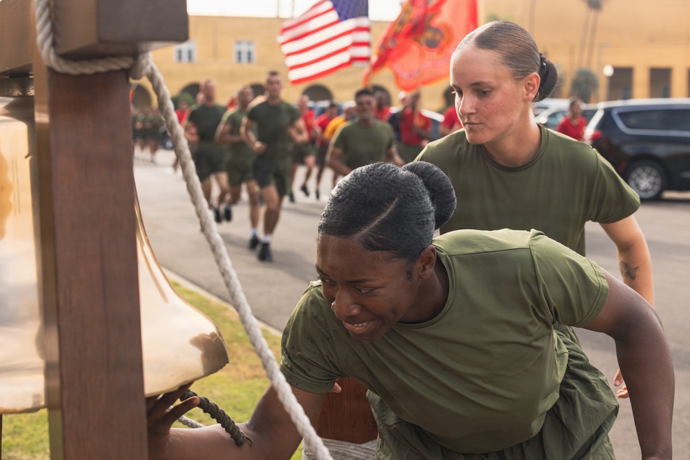 Two female service members in olive green military t-shirts are at the foreground, with one appearing to ring or touch a large bell while showing visible emotional strain. Behind them, a large formation of military personnel in matching uniforms are running in formation. American and Marine Corps flags are visible in the background, suggesting a military base setting.