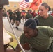 Two female service members in olive green military t-shirts are at the foreground, with one appearing to ring or touch a large bell while showing visible emotional strain. Behind them, a large formation of military personnel in matching uniforms are running in formation. American and Marine Corps flags are visible in the background, suggesting a military base setting.