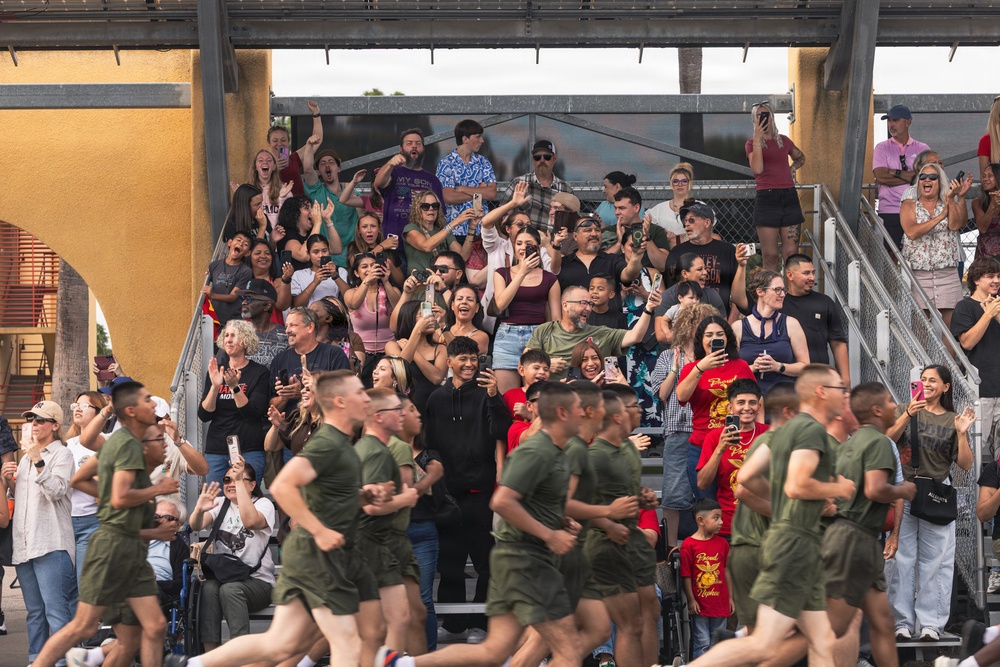 A large crowd of civilians cheers enthusiastically from bleachers as a formation of military recruits in olive green PT (physical training) uniforms runs past. The spectators, likely family members and supporters, are visibly emotional, clapping, waving, and recording with smartphones. The scene appears to be a military graduation or family day event, consistent with a Marine Corps recruit training ceremony.