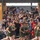 A large crowd of civilians cheers enthusiastically from bleachers as a formation of military recruits in olive green PT (physical training) uniforms runs past. The spectators, likely family members and supporters, are visibly emotional, clapping, waving, and recording with smartphones. The scene appears to be a military graduation or family day event, consistent with a Marine Corps recruit training ceremony.