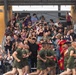 A large crowd of civilians cheers enthusiastically from bleachers as a formation of military recruits in olive green PT (physical training) uniforms runs past. The spectators, likely family members and supporters, are visibly emotional, clapping, waving, and recording with smartphones. The scene appears to be a military graduation or family day event, consistent with a Marine Corps recruit training ceremony.