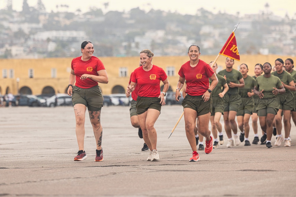 A group of female military personnel are running in formation on a large paved parade ground or drill field. Three individuals in red t-shirts and olive green shorts lead the formation, while a larger group in matching olive green PT uniforms follows behind. A guidon (unit flag) bearing the number '100' in U.S. Marine Corps colors (red and gold) is visible, suggesting this is a Marine Corps physical training run.