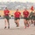 A group of female military personnel are running in formation on a large paved parade ground or drill field. Three individuals in red t-shirts and olive green shorts lead the formation, while a larger group in matching olive green PT uniforms follows behind. A guidon (unit flag) bearing the number '100' in U.S. Marine Corps colors (red and gold) is visible, suggesting this is a Marine Corps physical training run.