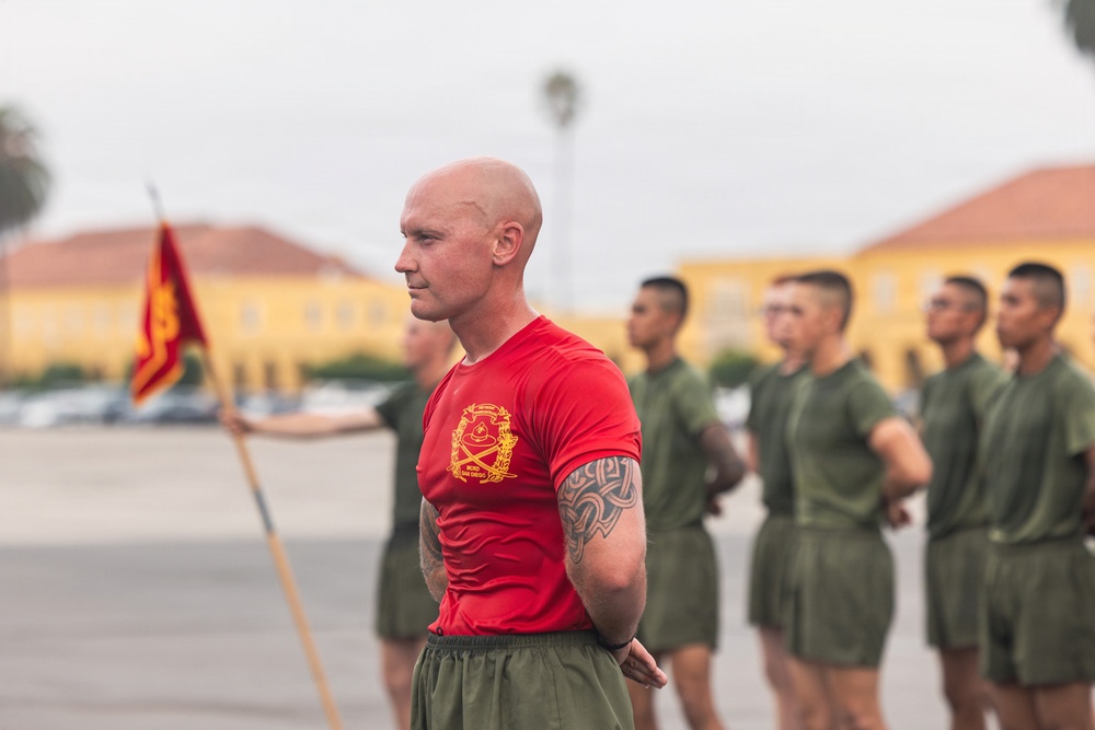 A bald male instructor or senior non-commissioned officer wearing a red t-shirt with a Marine Corps emblem stands in front of a formation of recruits or junior Marines in olive drab PT (physical training) uniforms. The setting appears to be a parade deck or drill field at what looks like Marine Corps Base Camp Pendleton or MCRD San Diego, identifiable by the characteristic yellow buildings in the background. A Marine Corps guidon flag is visible to the left.