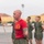 A bald male instructor or senior non-commissioned officer wearing a red t-shirt with a Marine Corps emblem stands in front of a formation of recruits or junior Marines in olive drab PT (physical training) uniforms. The setting appears to be a parade deck or drill field at what looks like Marine Corps Base Camp Pendleton or MCRD San Diego, identifiable by the characteristic yellow buildings in the background. A Marine Corps guidon flag is visible to the left.