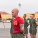 A bald male instructor or senior non-commissioned officer wearing a red t-shirt with a Marine Corps emblem stands in front of a formation of recruits or junior Marines in olive drab PT (physical training) uniforms. The setting appears to be a parade deck or drill field at what looks like Marine Corps Base Camp Pendleton or MCRD San Diego, identifiable by the characteristic yellow buildings in the background. A Marine Corps guidon flag is visible to the left.
