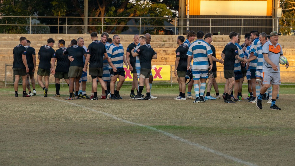 Two rugby teams are gathered on a grass field in a post-match handshake line, a traditional sportsmanship ritual. One team wears black jerseys with olive/khaki shorts, while the other wears blue and white hooped jerseys with matching shorts. The scene appears to be at dusk given the warm golden lighting, with concrete grandstand seating and advertising banners visible in the background.