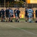 Two rugby teams are gathered on a grass field in a post-match handshake line, a traditional sportsmanship ritual. One team wears black jerseys with olive/khaki shorts, while the other wears blue and white hooped jerseys with matching shorts. The scene appears to be at dusk given the warm golden lighting, with concrete grandstand seating and advertising banners visible in the background.