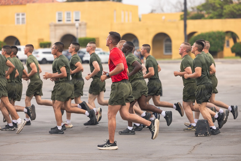 A large group of military personnel in olive green PT (physical training) uniforms are running in formation on a paved surface. One individual wearing a red shirt, likely an instructor or leader, runs among them in a central position. The setting appears to be a military base with a distinctive yellow Spanish-style building visible in the background.