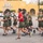 A large group of military personnel in olive green PT (physical training) uniforms are running in formation on a paved surface. One individual wearing a red shirt, likely an instructor or leader, runs among them in a central position. The setting appears to be a military base with a distinctive yellow Spanish-style building visible in the background.