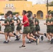 A large group of military personnel in olive green PT (physical training) uniforms are running in formation on a paved surface. One individual wearing a red shirt, likely an instructor or leader, runs among them in a central position. The setting appears to be a military base with a distinctive yellow Spanish-style building visible in the background.