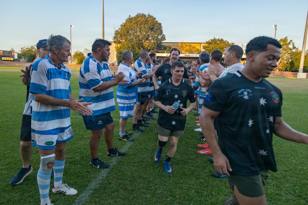 A rugby match post-game handshake ceremony is taking place on a grass field, with two teams forming a tunnel or line to congratulate each other. One team wears blue and white hooped jerseys while the opposing team wears black jerseys featuring star motifs resembling the Australian flag design. The setting appears to be an outdoor rugby ground during late afternoon or early evening light.