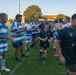 A rugby match post-game handshake ceremony is taking place on a grass field, with two teams forming a tunnel or line to congratulate each other. One team wears blue and white hooped jerseys while the opposing team wears black jerseys featuring star motifs resembling the Australian flag design. The setting appears to be an outdoor rugby ground during late afternoon or early evening light.