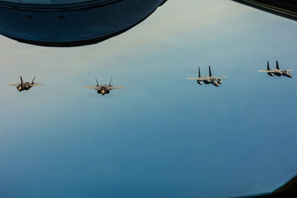 Four military jet aircraft are flying in a loose formation over what appears to be a large body of water, photographed from the window or observation port of another aircraft. The formation consists of two F-35 Lightning II stealth fighters on the left and two F-15 Eagle fighters on the right. The image captures an aerial refueling or escort mission scenario with clear blue sky and water visible below.