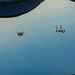 Four military jet aircraft are flying in a loose formation over what appears to be a large body of water, photographed from the window or observation port of another aircraft. The formation consists of two F-35 Lightning II stealth fighters on the left and two F-15 Eagle fighters on the right. The image captures an aerial refueling or escort mission scenario with clear blue sky and water visible below.