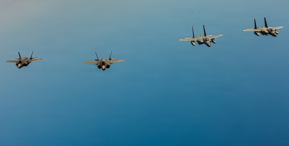 Four military jet aircraft are flying in a loose formation against a clear blue sky. The formation consists of two distinct aircraft types: two F-35 Lightning II stealth fighters on the left and two F-15 Eagle fighters on the right. The image appears to be an aerial photograph taken from another aircraft at a similar altitude.