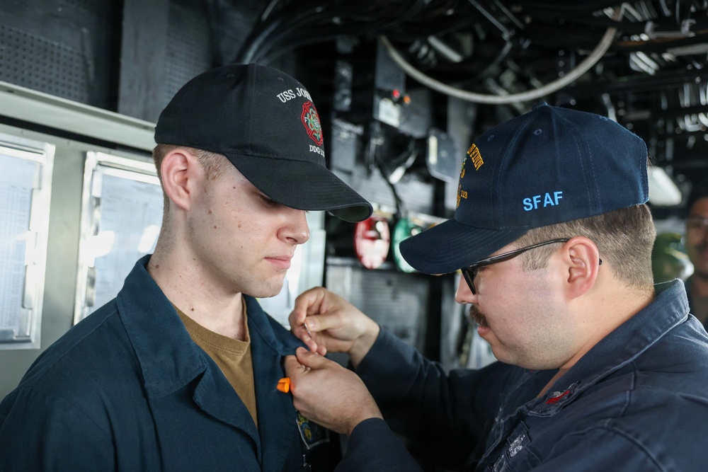 Two U.S. Navy sailors are shown aboard what appears to be a naval vessel, with one sailor pinning something onto the collar or chest area of the other. The sailor on the right is wearing a cap with 'SFAF' visible in yellow lettering, and both wear dark navy working uniforms. This appears to be a military ceremony, likely a promotion or qualification pinning event.