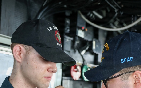 Two U.S. Navy sailors are shown aboard what appears to be a naval vessel, with one sailor pinning something onto the collar or chest area of the other. The sailor on the right is wearing a cap with 'SFAF' visible in yellow lettering, and both wear dark navy working uniforms. This appears to be a military ceremony, likely a promotion or qualification pinning event.