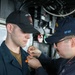 Two U.S. Navy sailors are shown aboard what appears to be a naval vessel, with one sailor pinning something onto the collar or chest area of the other. The sailor on the right is wearing a cap with 'SFAF' visible in yellow lettering, and both wear dark navy working uniforms. This appears to be a military ceremony, likely a promotion or qualification pinning event.