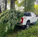A white Ford F-250 FX4 pickup truck is parked in a wooded, grassy area with its truck bed loaded with large cut plant stalks with broad leaves overhanging the tailgate. In the background, a second vehicle with visible blue emergency lights is present. The scene appears to be an outdoor law enforcement operation based on the presence of the emergency vehicle.