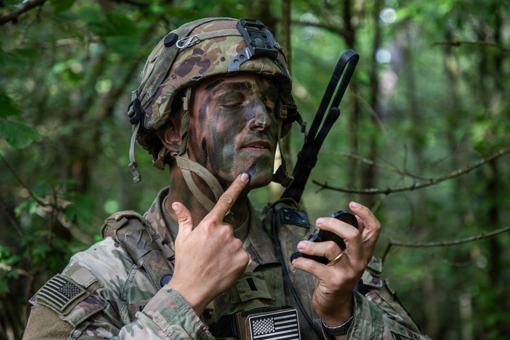 A U.S. Army soldier in full combat gear is operating a handheld radio in a forested environment. The soldier is wearing OCP (Operational Camouflage Pattern) uniform and helmet with face camouflage paint applied. He appears to be communicating or adjusting radio settings during what appears to be a field training exercise.