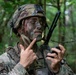A U.S. Army soldier in full combat gear is operating a handheld radio in a forested environment. The soldier is wearing OCP (Operational Camouflage Pattern) uniform and helmet with face camouflage paint applied. He appears to be communicating or adjusting radio settings during what appears to be a field training exercise.
