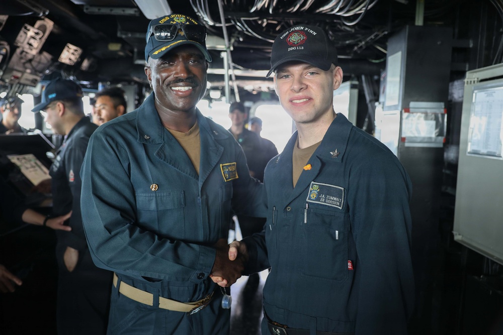 Two U.S. Navy sailors are shaking hands aboard what appears to be the bridge or interior of USS John Finn (DDG-113), a guided-missile destroyer. The sailor on the right wears a name tag reading 'J.A. Zumwalt' and is assigned to 'ER Division,' while both wear USS John Finn DDG-113 caps. Several other crew members are visible in the background, suggesting a formal recognition or ceremony moment.
