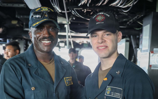 Two U.S. Navy sailors are shaking hands aboard what appears to be the bridge or interior of USS John Finn (DDG-113), a guided-missile destroyer. The sailor on the right wears a name tag reading 'J.A. Zumwalt' and is assigned to 'ER Division,' while both wear USS John Finn DDG-113 caps. Several other crew members are visible in the background, suggesting a formal recognition or ceremony moment.