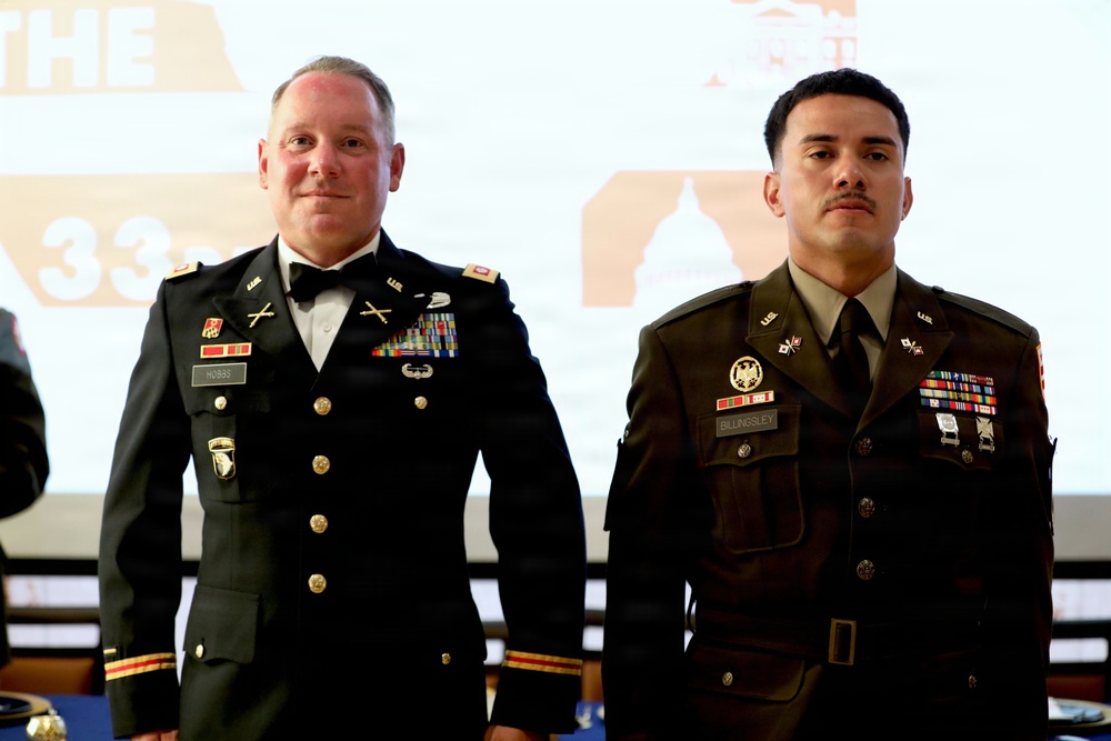 Two U.S. Army soldiers in dress uniforms are standing side by side at what appears to be a formal military ceremony or ball. The soldier on the left wears the Army Service Uniform (ASU) in black with a bow tie, while the soldier on the right wears the Army Green Service Uniform. Both display multiple ribbons, badges, and name tags visible on their uniforms.
