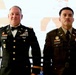 Two U.S. Army soldiers in dress uniforms are standing side by side at what appears to be a formal military ceremony or ball. The soldier on the left wears the Army Service Uniform (ASU) in black with a bow tie, while the soldier on the right wears the Army Green Service Uniform. Both display multiple ribbons, badges, and name tags visible on their uniforms.
