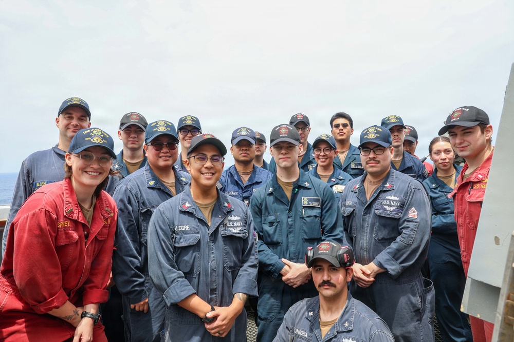 A group of approximately 20 U.S. Navy personnel posing for a group photo on the deck of a naval vessel at sea. Most are wearing navy blue coveralls and matching ship-assigned caps, with one individual in red coveralls on the left. The group appears to be in good spirits, smiling for the camera.