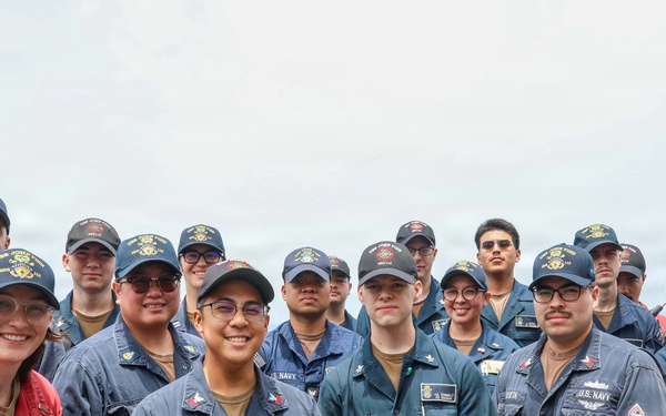 A group of approximately 20 U.S. Navy personnel posing for a group photo on the deck of a naval vessel at sea. Most are wearing navy blue coveralls and matching ship-assigned caps, with one individual in red coveralls on the left. The group appears to be in good spirits, smiling for the camera.