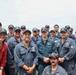 A group of approximately 20 U.S. Navy personnel posing for a group photo on the deck of a naval vessel at sea. Most are wearing navy blue coveralls and matching ship-assigned caps, with one individual in red coveralls on the left. The group appears to be in good spirits, smiling for the camera.