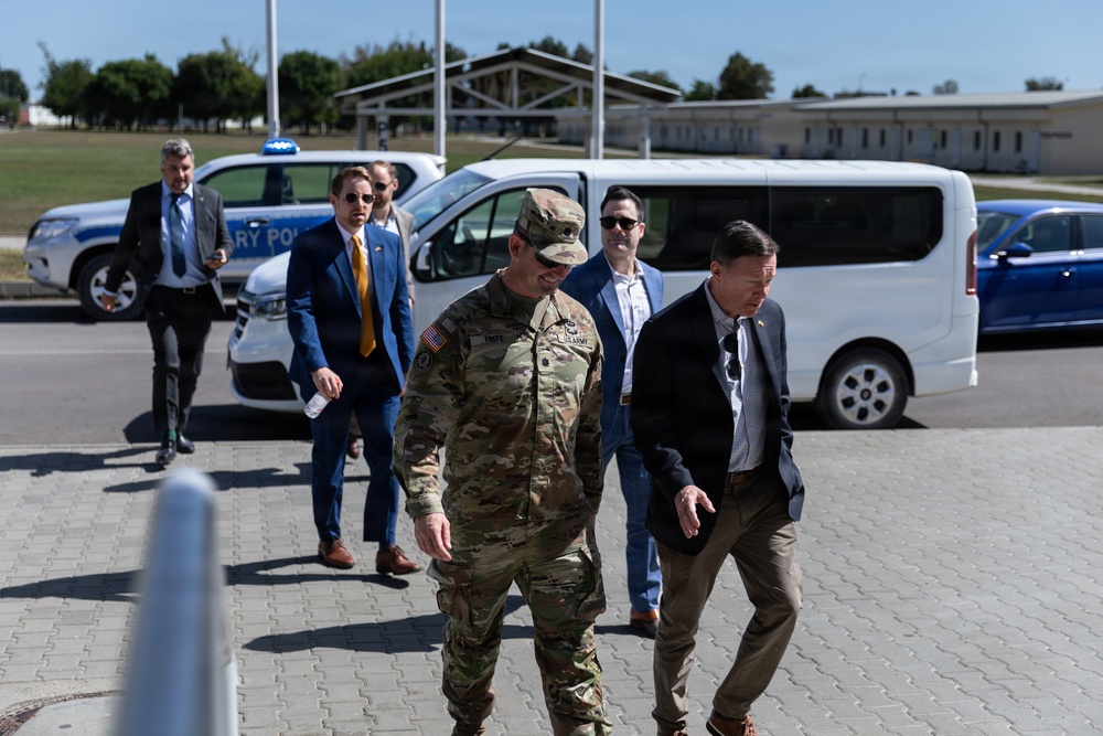 A group of individuals, including a U.S. Army soldier in multicam uniform and several civilians in business attire, are walking across a paved area at what appears to be a military installation. A Military Police vehicle is visible in the background along with a white passenger van. The setting suggests an official visit or delegation tour at a military base.