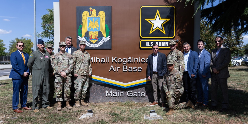 A group of military personnel and civilians pose for a formal photograph in front of the Mihail Kogălniceanu Air Base Main Gate sign in Romania. The sign displays both the Romanian military coat of arms and the U.S. Army logo, indicating a joint or bilateral military presence. The setting appears to be an official visit or ceremony at the base entrance.