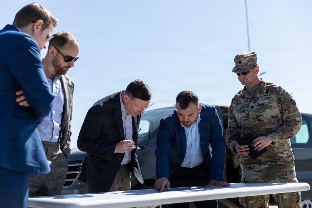 A group of five men are gathered outdoors around a white folding table, reviewing documents or plans. The group includes four civilians in business attire and one U.S. Army soldier in OCP (Operational Camouflage Pattern) uniform. The setting appears to be an outdoor briefing or site visit, likely on or near a military installation.