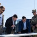 A group of five men are gathered outdoors around a white folding table, reviewing documents or plans. The group includes four civilians in business attire and one U.S. Army soldier in OCP (Operational Camouflage Pattern) uniform. The setting appears to be an outdoor briefing or site visit, likely on or near a military installation.