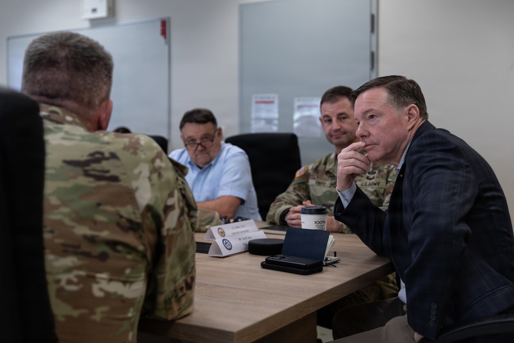 A formal meeting or briefing is taking place at a conference table involving military personnel in U.S. Army camouflage uniforms and civilians in business attire. The group appears engaged in a discussion, with one civilian leaning forward attentively. Name placards and a coffee cup are visible on the table, suggesting an official or structured setting.