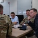 A formal meeting or briefing is taking place at a conference table involving military personnel in U.S. Army camouflage uniforms and civilians in business attire. The group appears engaged in a discussion, with one civilian leaning forward attentively. Name placards and a coffee cup are visible on the table, suggesting an official or structured setting.