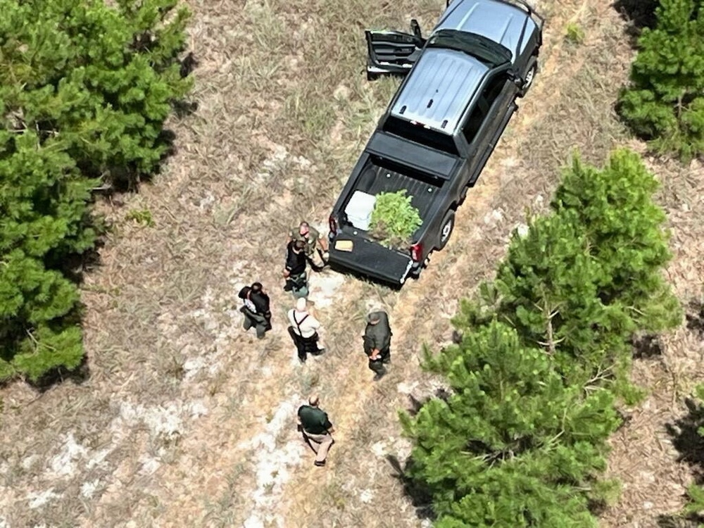 An aerial photograph shows several individuals gathered around a black pickup truck in a rural, wooded clearing. The truck's bed appears to contain green plant material, and multiple people wearing what appear to be law enforcement or tactical uniforms are present at the scene. The image appears to document an outdoor law enforcement operation.