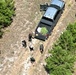 An aerial photograph shows several individuals gathered around a black pickup truck in a rural, wooded clearing. The truck's bed appears to contain green plant material, and multiple people wearing what appear to be law enforcement or tactical uniforms are present at the scene. The image appears to document an outdoor law enforcement operation.