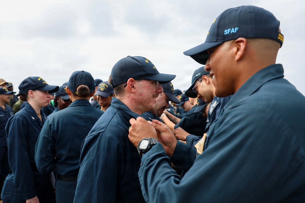A group of military personnel in dark navy blue uniforms and caps are gathered outdoors, likely during an inspection or ceremony. A senior member wearing a cap marked 'SFAF' appears to be pinning something onto or inspecting the collar/chest area of another service member. Multiple uniformed individuals are visible in the background under a partly cloudy sky.
