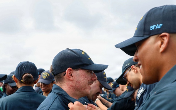 A group of military personnel in dark navy blue uniforms and caps are gathered outdoors, likely during an inspection or ceremony. A senior member wearing a cap marked 'SFAF' appears to be pinning something onto or inspecting the collar/chest area of another service member. Multiple uniformed individuals are visible in the background under a partly cloudy sky.