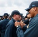 A group of military personnel in dark navy blue uniforms and caps are gathered outdoors, likely during an inspection or ceremony. A senior member wearing a cap marked 'SFAF' appears to be pinning something onto or inspecting the collar/chest area of another service member. Multiple uniformed individuals are visible in the background under a partly cloudy sky.