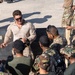 A military instructor in desert-pattern combat shirt and sunglasses appears to be conducting a briefing or demonstration for a multinational group of soldiers. The gathering includes personnel wearing various camouflage patterns and insignia, suggesting a joint or coalition training exercise. The setting appears to be an outdoor hardstand or port facility.