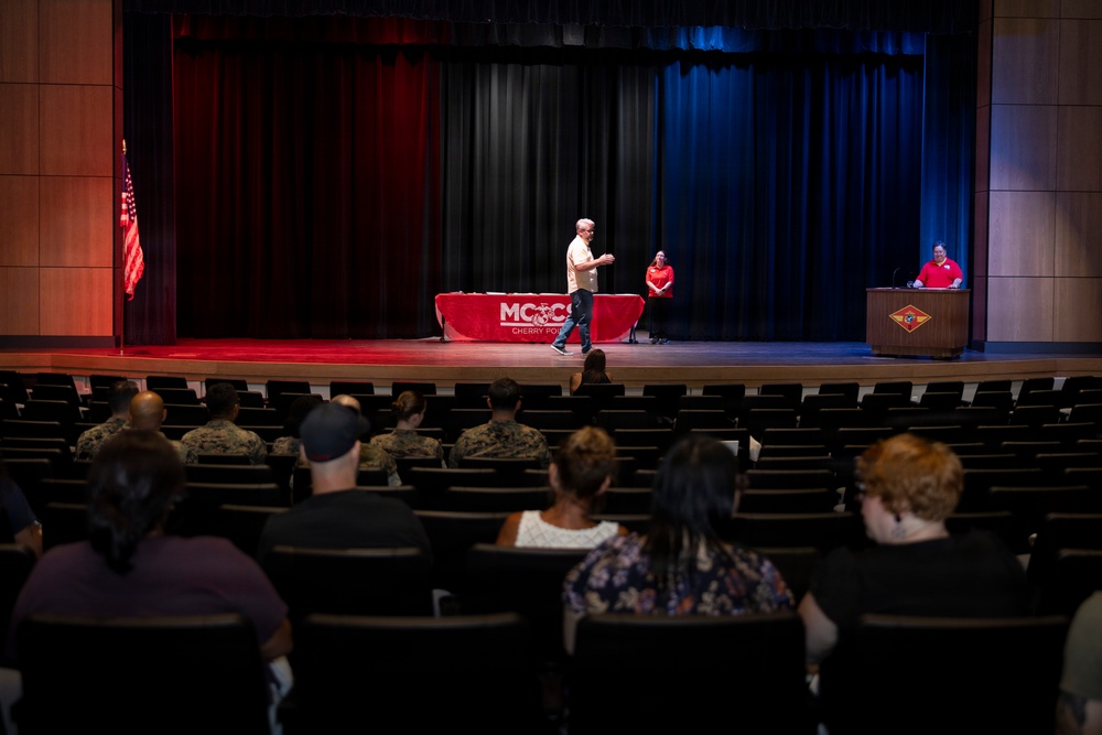 An event is taking place in a theater or auditorium at MCCS (Marine Corps Community Services) Cherry Point, as indicated by the red-draped table on stage. A man in civilian clothes is walking across the stage while two other individuals in red shirts are positioned at the table and podium. The audience includes a mix of military personnel in camouflage uniforms and civilians seated in theater chairs.