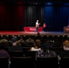 An event is taking place in a theater or auditorium at MCCS (Marine Corps Community Services) Cherry Point, as indicated by the red-draped table on stage. A man in civilian clothes is walking across the stage while two other individuals in red shirts are positioned at the table and podium. The audience includes a mix of military personnel in camouflage uniforms and civilians seated in theater chairs.
