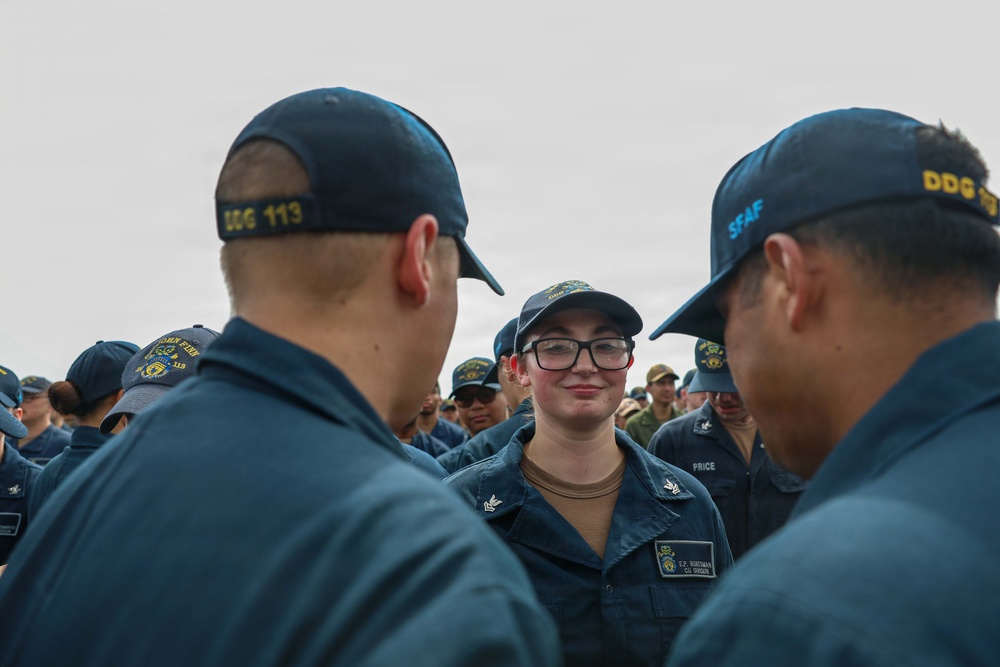 A group of U.S. Navy sailors in dark blue working uniforms are gathered together, with one female sailor wearing glasses and a name tag reading 'PRICE' visible in the center foreground. The sailors are wearing caps with unit designations including 'DDG 113' and 'SFAF' visible on the back of caps in the foreground. The scene appears to be an outdoor formation or informal gathering involving multiple personnel.