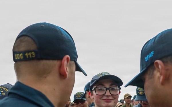 A group of U.S. Navy sailors in dark blue working uniforms are gathered together, with one female sailor wearing glasses and a name tag reading 'PRICE' visible in the center foreground. The sailors are wearing caps with unit designations including 'DDG 113' and 'SFAF' visible on the back of caps in the foreground. The scene appears to be an outdoor formation or informal gathering involving multiple personnel.